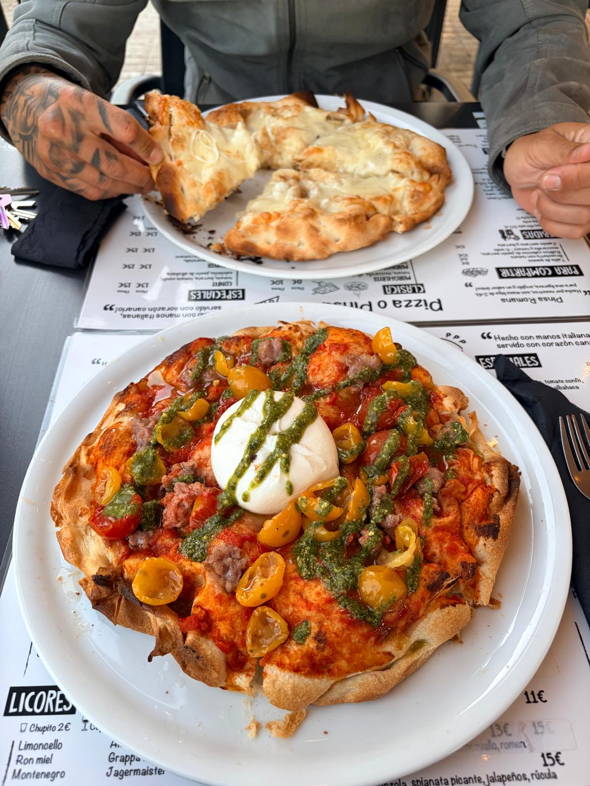 Pizza con burrata, pesto, tomates cherry amarillos y salchicha; detrás, una pizza de queso